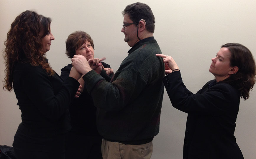 Four adults stand in a circle, each person pointing at another. Two women point at a man in glasses, and he points at a woman. Another woman points at him from behind. All appear focused and serious.