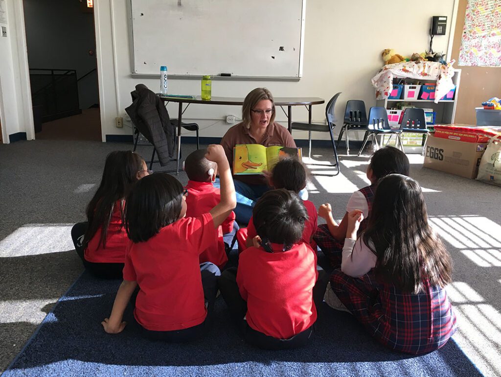 A woman reads a picture book to a group of young children sitting on the floor in a classroom. The children, some raising their hands, are wearing red shirts and uniforms. Sunlight streams through the windows.