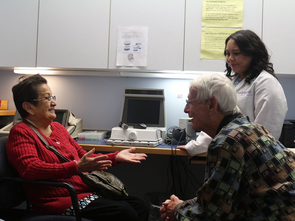 Two older adults sit and talk with a healthcare professional in an exam room. The woman in red gestures with her hands while the man and the professional listen, creating a warm, engaged atmosphere. Medical equipment is visible in the background.
