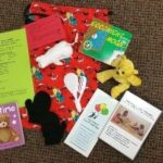 A collection of bedtime-themed items on a carpet: two children's books, two plush toys, a white mouse toy, a small racket, animal cutouts, printed activity guides, and a red drawstring bag with cartoon characters.