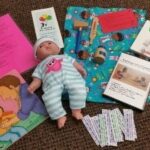 A collection of children's doctor-themed items on a carpet: a baby doll in striped clothes, books, a stethoscope toy, a mallet, a syringe, paper labels, activity sheets, and a colorful blue cloth with cartoon prints.