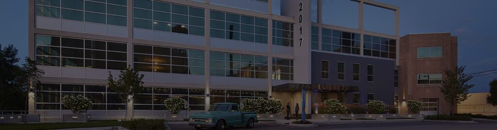 A modern office building with large glass windows at dusk, featuring a vintage green pickup truck parked in front and trees and shrubs lining the sidewalk. The building's address, 20111, is displayed vertically on a column.