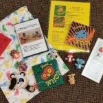 A set of children's items on a carpet, including a patterned drawstring bag, small plush toys, a soft "Peek-a-Who?" book, activity cards, and an information pamphlet with balloons on the cover.