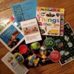 Toys, board books about vehicles, a road map mat, toy cars, and informational papers are spread out on a wooden floor, arranged for a child's activity or learning session about things that go.
